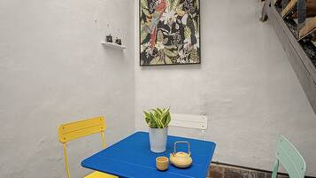 A small outdoor seating area with a blue table, yellow and green chairs, a potted plant, and a teapot.