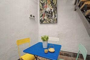 Un petit espace extérieur aménagé avec une table bleue, des chaises jaunes et vertes, une plante en pot et une théière.