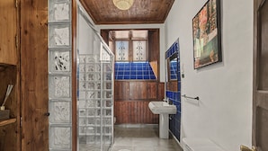 Un pasillo estrecho con una puerta de madera, una puerta de vidrio con diseños decorativos, una pared de azulejos azules, un lavamanos blanco y un cuadro enmarcado en la pared.