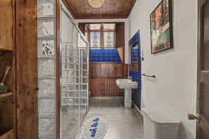 Un pasillo estrecho con una puerta de madera, una puerta de vidrio con diseños decorativos, una pared de azulejos azules, un lavamanos blanco y un cuadro enmarcado en la pared.