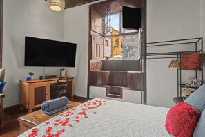 Un dormitorio con cama, televisión, cómoda de madera y ventana con vistas a edificios.