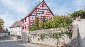 Un edificio tradicional de entramado de madera con techo inclinado, pared de piedra y cerca de madera, bajo un cielo despejado.