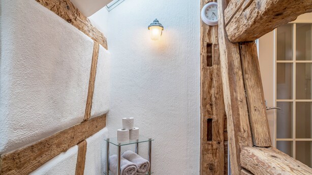 Un baño estrecho con vigas de madera, un toallero de vidrio con toallas y una lámpara de pared.