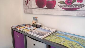 Un comptoir d’accueil avec un présentoir à magazines, un tableau représentant des fruits et une petite lampe de bureau.