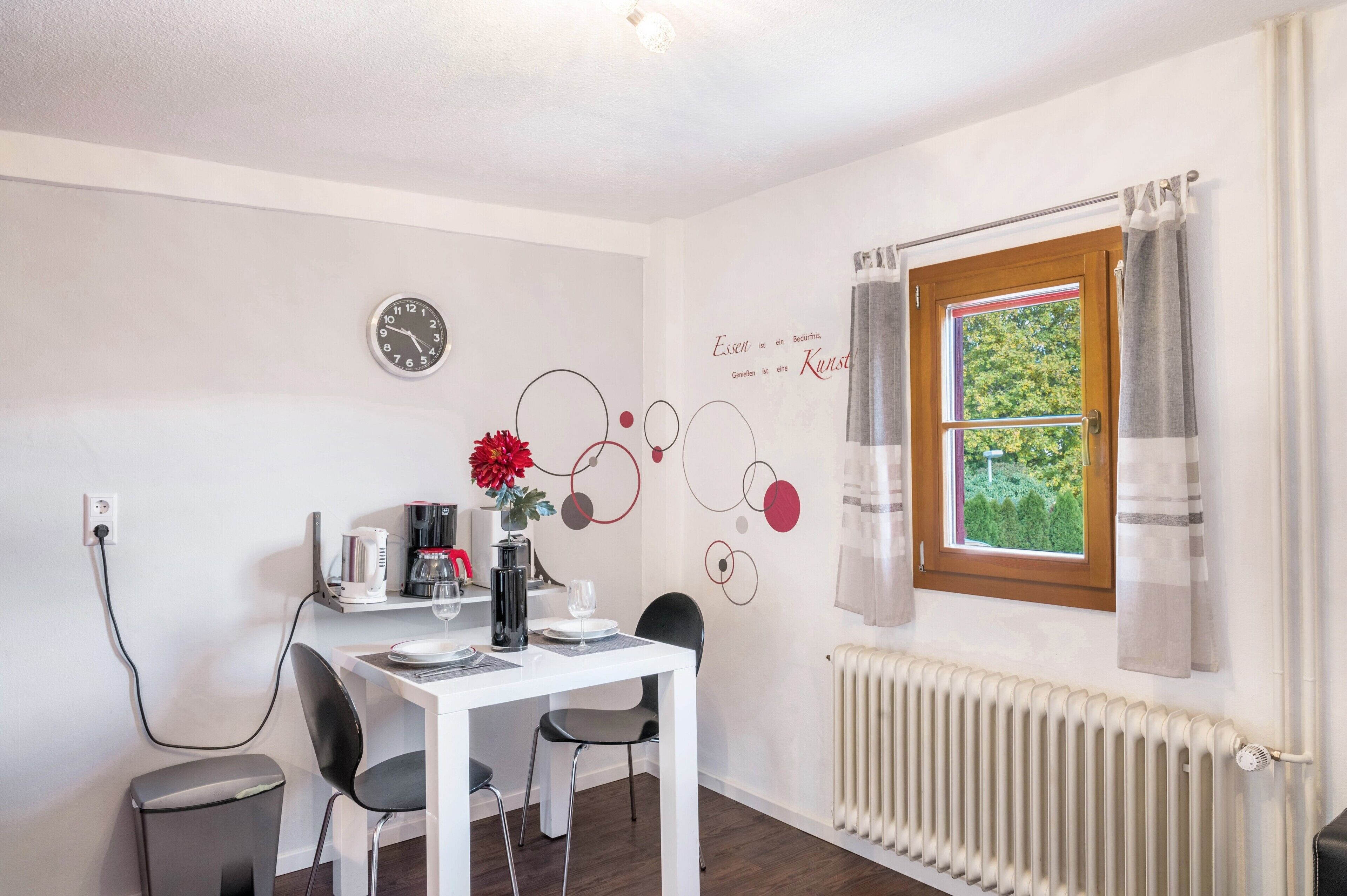Un coin repas avec une table blanche, des chaises noires, une horloge, un radiateur et une fenêtre avec des rideaux.