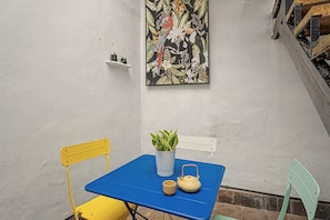 Un petit espace extérieur aménagé avec une table bleue, des chaises jaunes et vertes, une plante en pot et une théière.