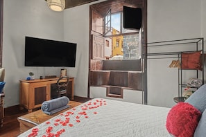 A bedroom with a bed, a television, a wooden dresser, and a window with a view of buildings.