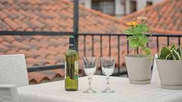 Una mesa con una botella de vino, dos copas, una planta en maceta con flores amarillas y una suculenta en maceta en un balcón con tejados de cerámica al fondo.