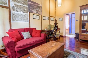 A red sofa with a patterned pillow, a wooden coffee table, a dining table with chairs, framed pictures on the wall, and a staircase leading to another room.