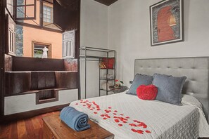 Une chambre à coucher avec un grand lit, une tête de lit, une table de chevet, une étagère avec des vêtements pliés et une fenêtre donnant sur un immeuble.