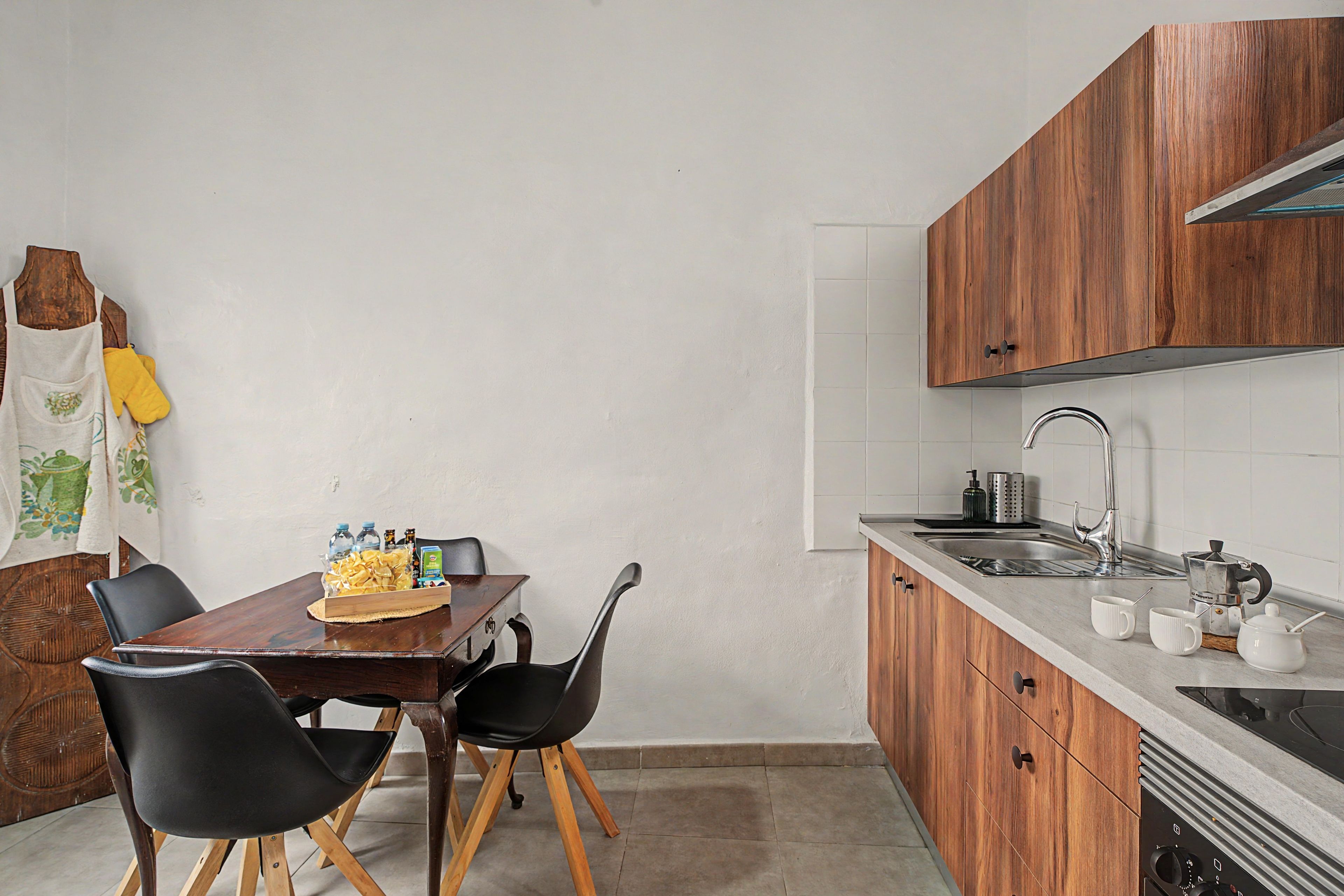 Een kleine keuken met houten kastjes, een spoelbak en een eethoek met twee stoelen.
