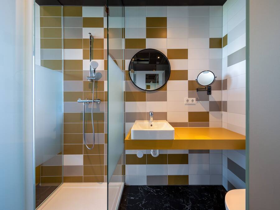Un baño moderno con cabina de ducha de vidrio, un lavamanos blanco con encimera amarilla y un espejo redondo en la pared.