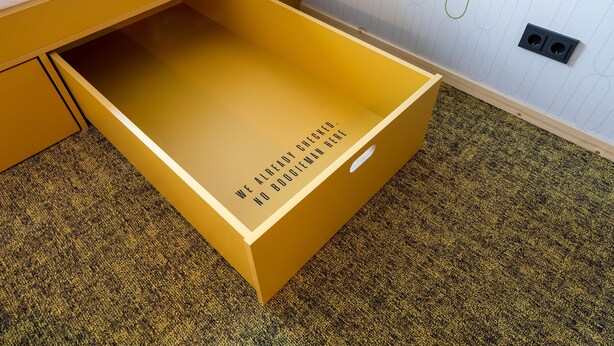 A yellow drawer with text on the inside, a bed with a white mattress, and a patterned wall. - Mr Jigs (Venlo)