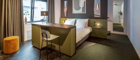 A hotel room with a bed, a desk, a chair, a small stool, and a window with curtains.