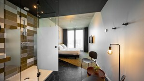 Een moderne hotelkamer met een glazen douchecabine, een bed met uitzicht, een zithoek en een kleine tafel met een plantje.