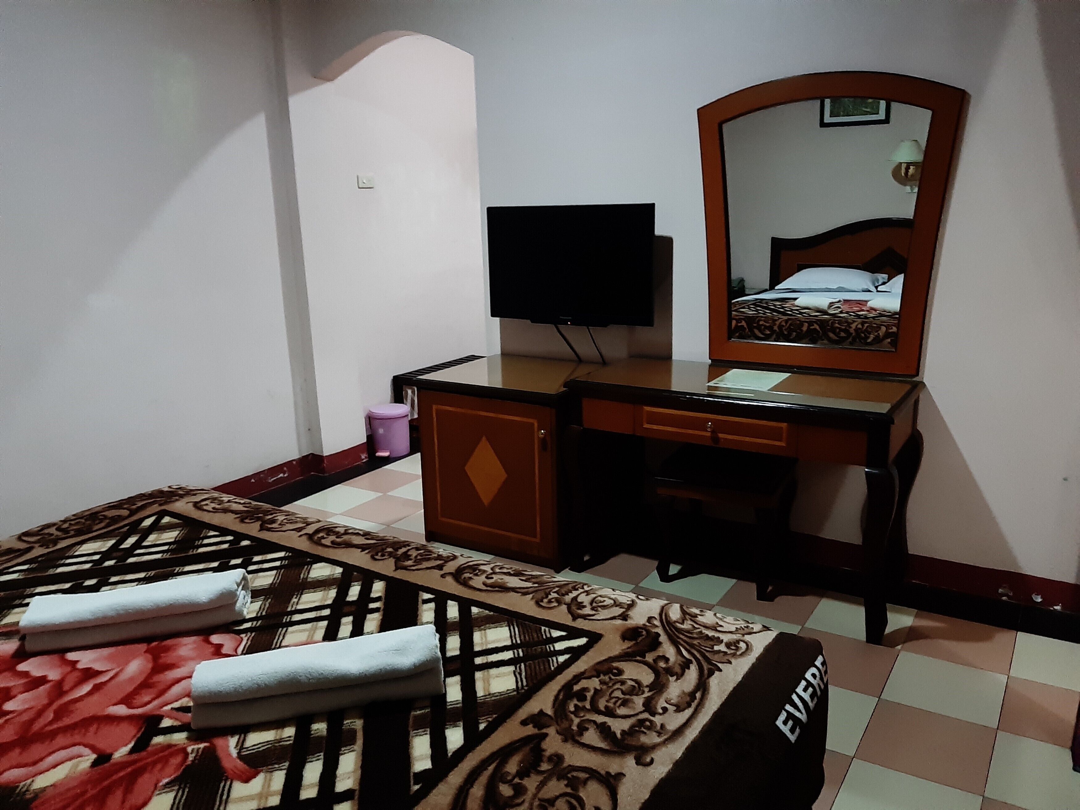 Una habitación de hotel con una cama, un escritorio con espejo, un televisor y un pequeño cubo de basura.