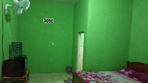 A small room with green walls, a bed with a floral bedspread, a desk with a chair, a water dispenser, and a ceiling fan. - Pondok Sundawa (Pangandaran)