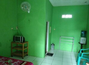 A room with green walls, a bed with a floral bedspread, a small wooden shelf with a TV, a standing fan, a water dispenser, and a metal rack. - Pondok Sundawa (Pangandaran)