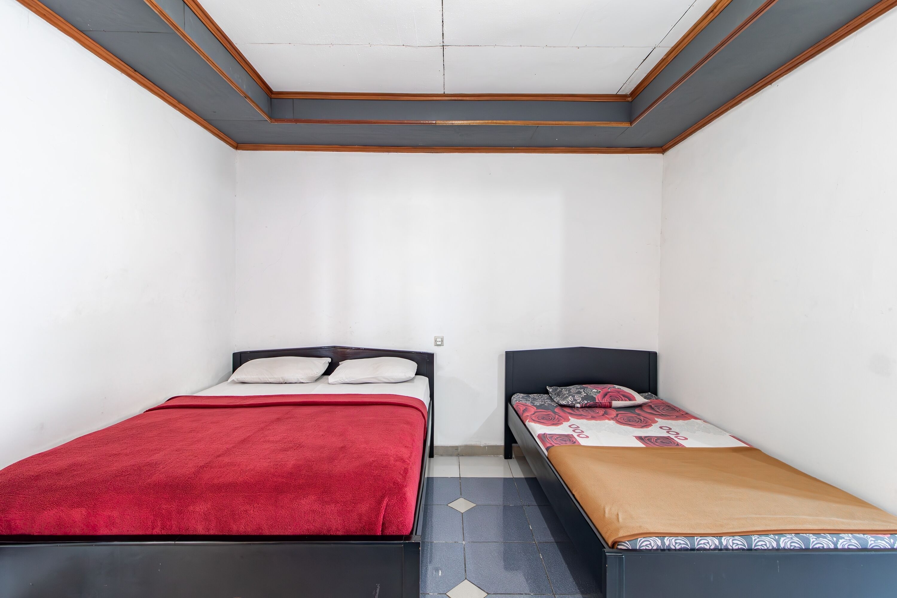Une petite pièce bien éclairée, avec deux lits : l’un est recouvert d’une couverture rouge et l’autre d’une couvre-lit à motifs.