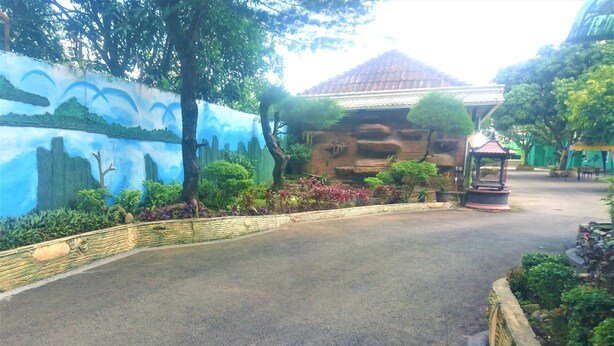 A paved pathway leading to a building with a tiled roof, surrounded by lush greenery and a painted mural. - Hotel Bukit Panembahan (Durin Simbelang)