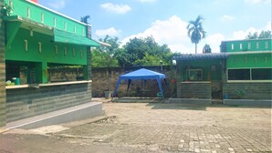A courtyard with a blue tent, green buildings, and a palm tree. - Hotel Bukit Panembahan (Durin Simbelang)