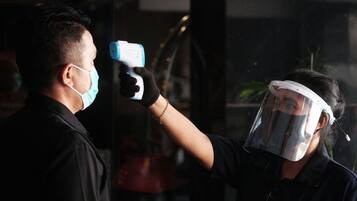 A person wearing a face shield and gloves, holding a device near another person's face.