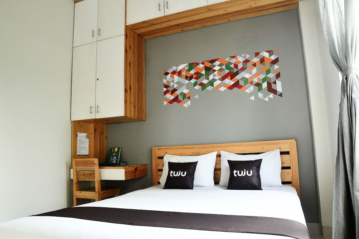 A bedroom with a wooden headboard, a bed with white bedding, two black pillows with 'tuju' text, a geometric wall art piece, and a wooden chair.