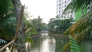 A calm water feature with a bridge, surrounded by lush greenery and a tall building in the background. - Apartment Green Lake View Ciputat by Celebrity Room (South Tangerang)