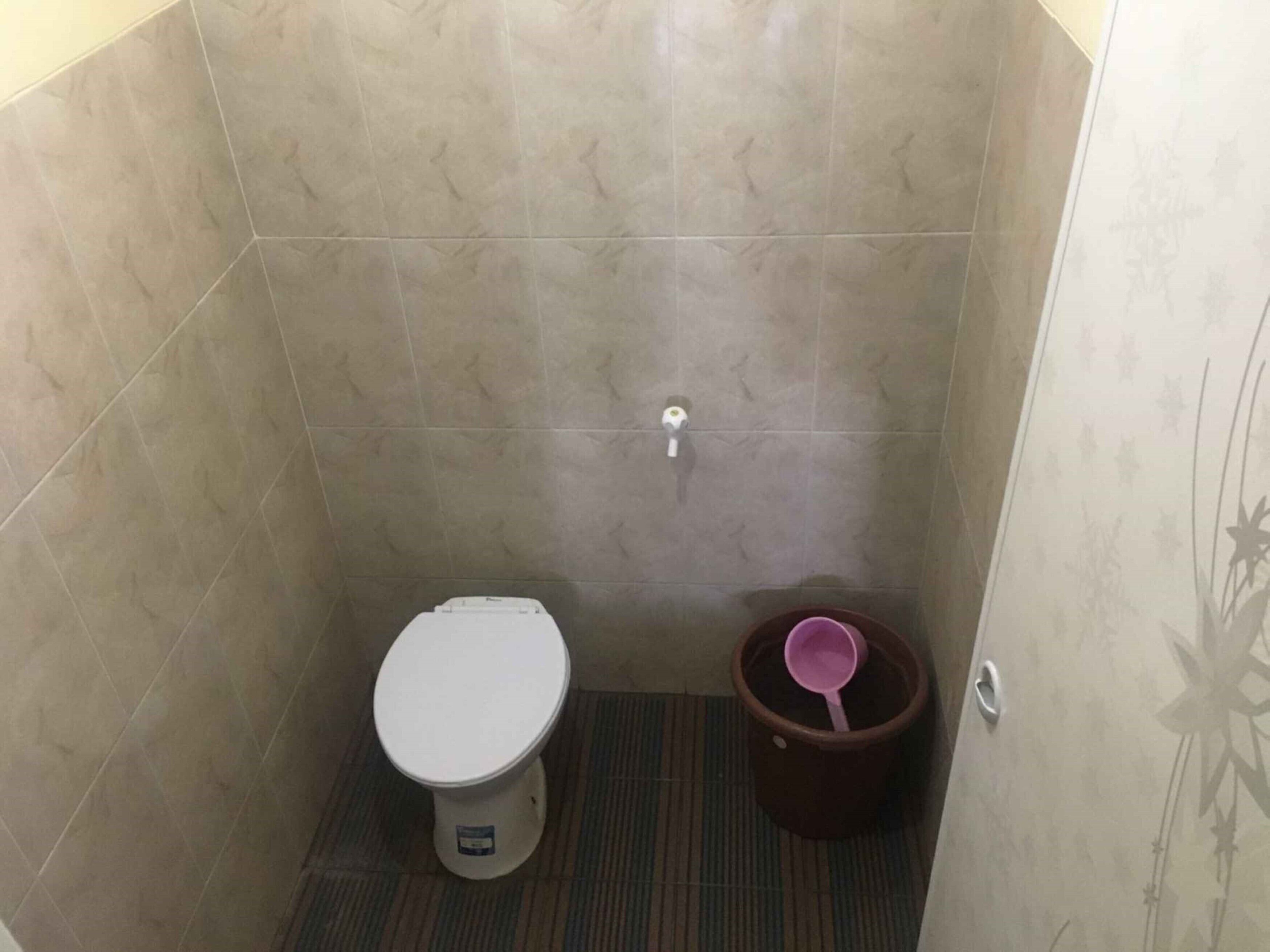 A small, tiled bathroom with a toilet, a trash bin, and a showerhead on the wall.