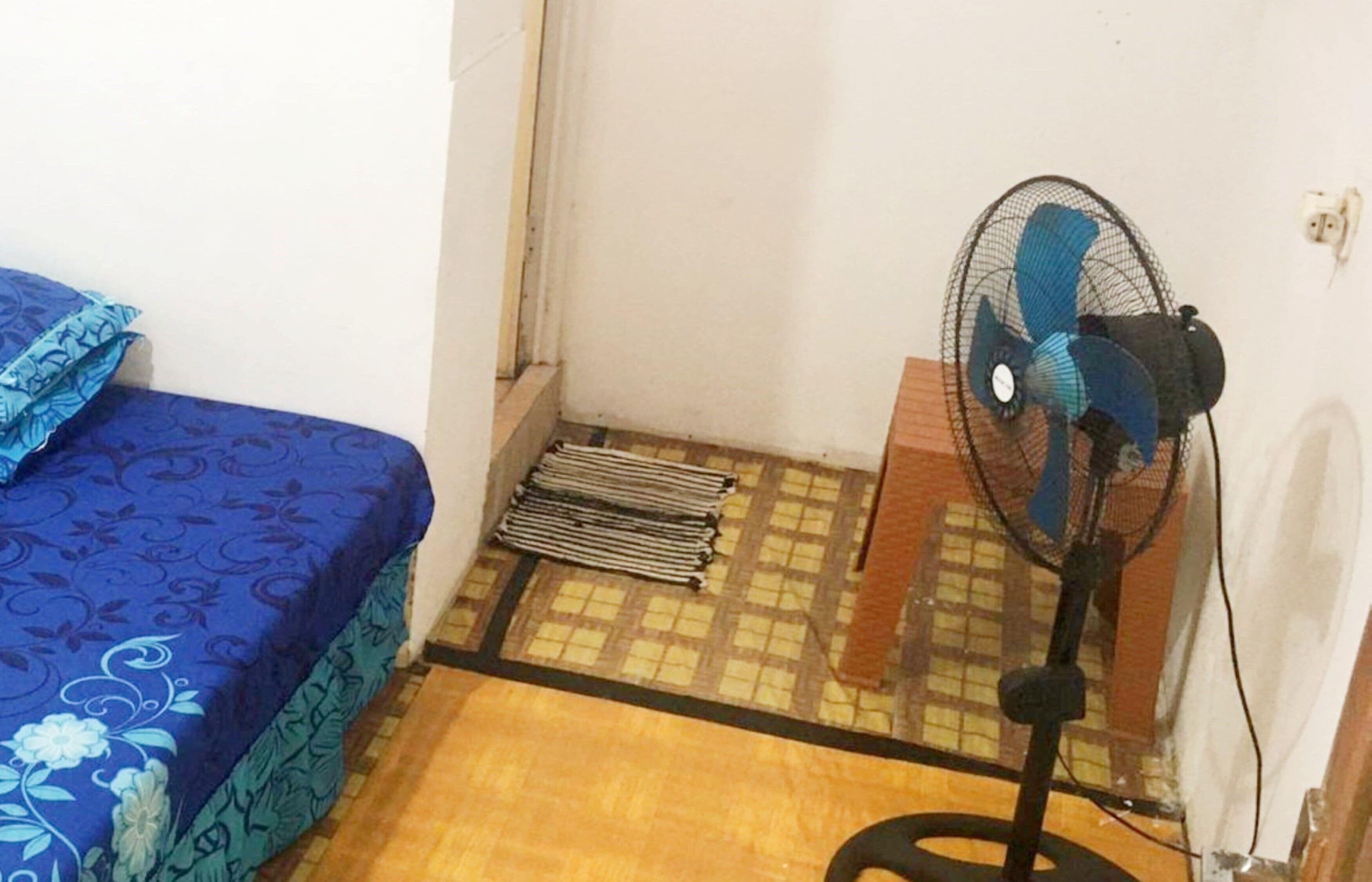 A small room with a bed, a standing fan, and a radiator.