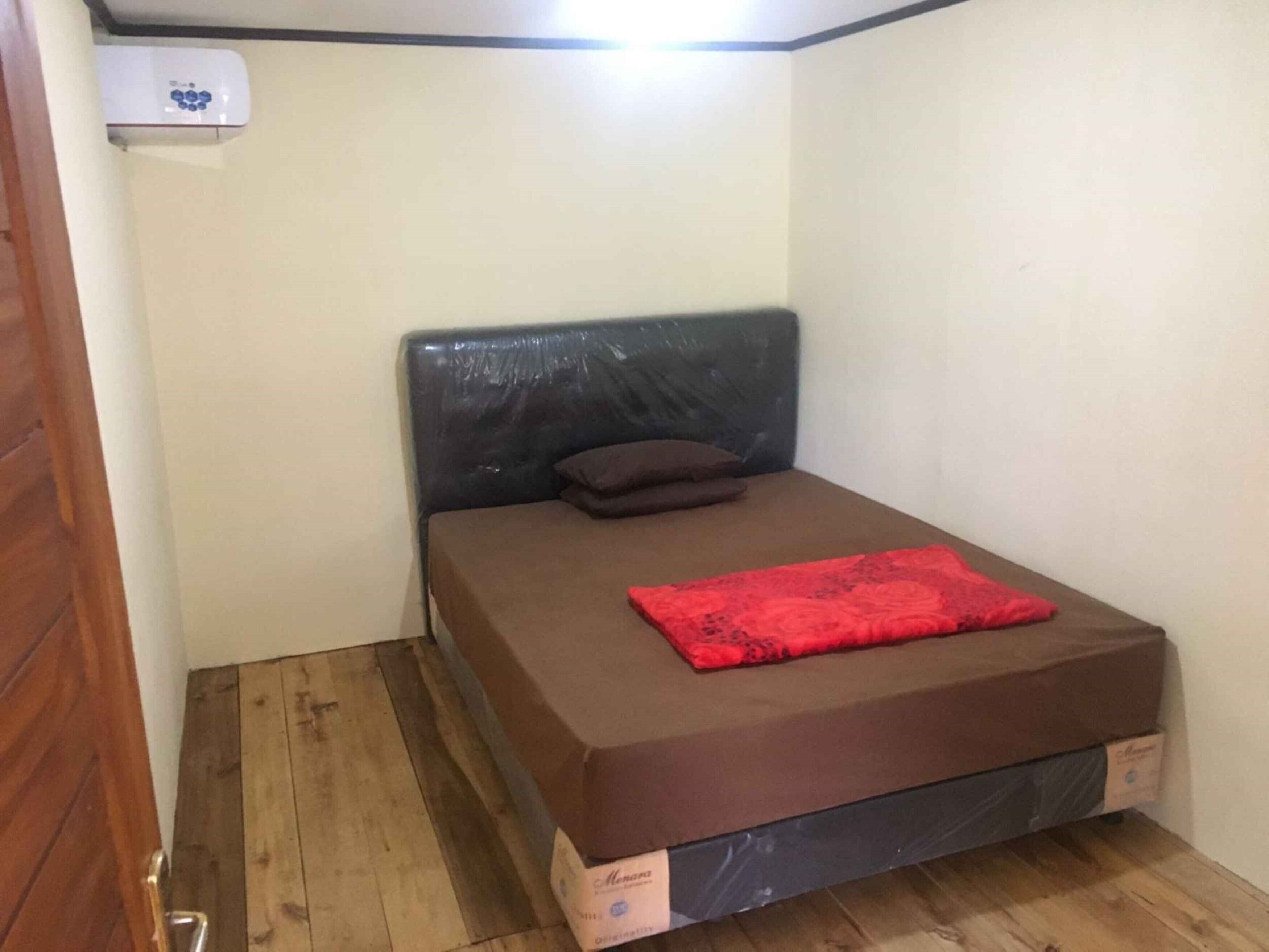 A small, single-bed room with a brown mattress, a black headboard, and a red blanket. An air conditioning unit is mounted on the wall above the bed.