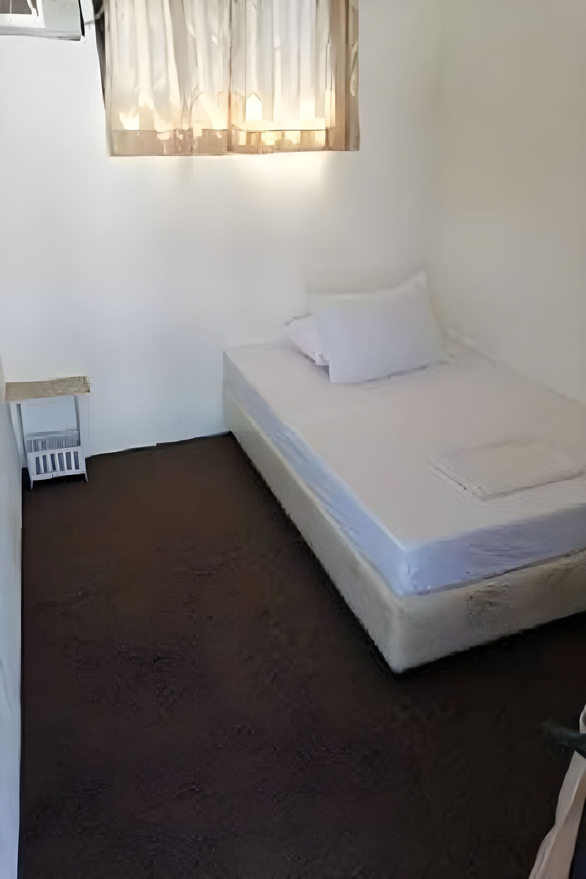 Una habitación pequeña con cama individual, colchón blanco, una mesita de noche y una ventana con cortinas translúcidas.