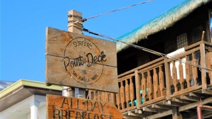 Ein Holzschild mit der Aufschrift 'Surfer's Point Deck' und 'All Day Breakfast', befestigt an einem Pfosten.
