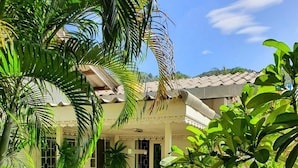 A covered outdoor seating area with benches and a table, surrounded by lush greenery and a building with a tiled roof. - Muchshima House (Choeng Thale)