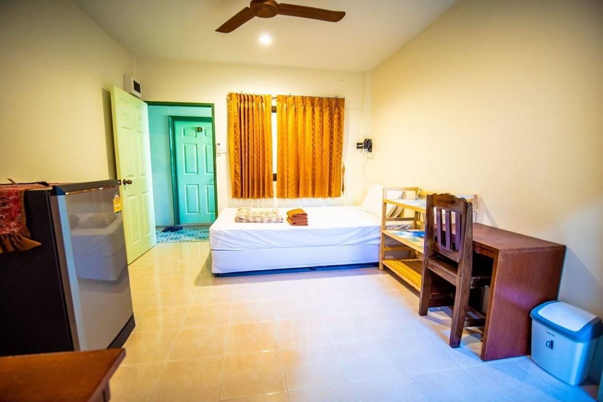 A bedroom with a bed, a ceiling fan, a window with curtains, a wooden chair, a small table, and a trash bin.