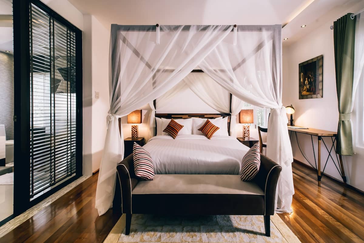 A bedroom with a four-poster bed, bedside lamps, a nightstand, a wooden floor, and a view through a window with blinds.