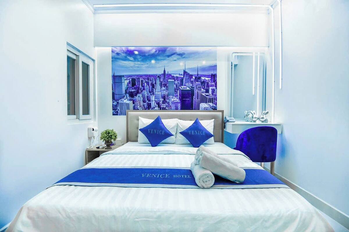 Une chambre d’hôtel avec un lit orné d’une fresque représentant un paysage urbain, deux oreillers avec le mot « Venise » inscrit dessus, une couverture bleue et une serviette sur le lit.