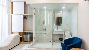Un baño moderno con ducha de vidrio, lavamanos blanco y un inodoro. Hay una silla azul y una mesita con una caja de pañuelos.