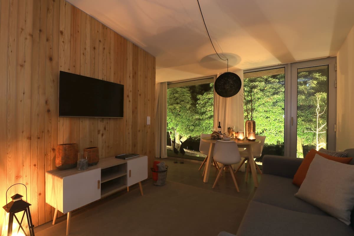 A modern living room with a wooden wall, a flat-screen TV, a dining area with a table and chairs, and a large window with a view of greenery.