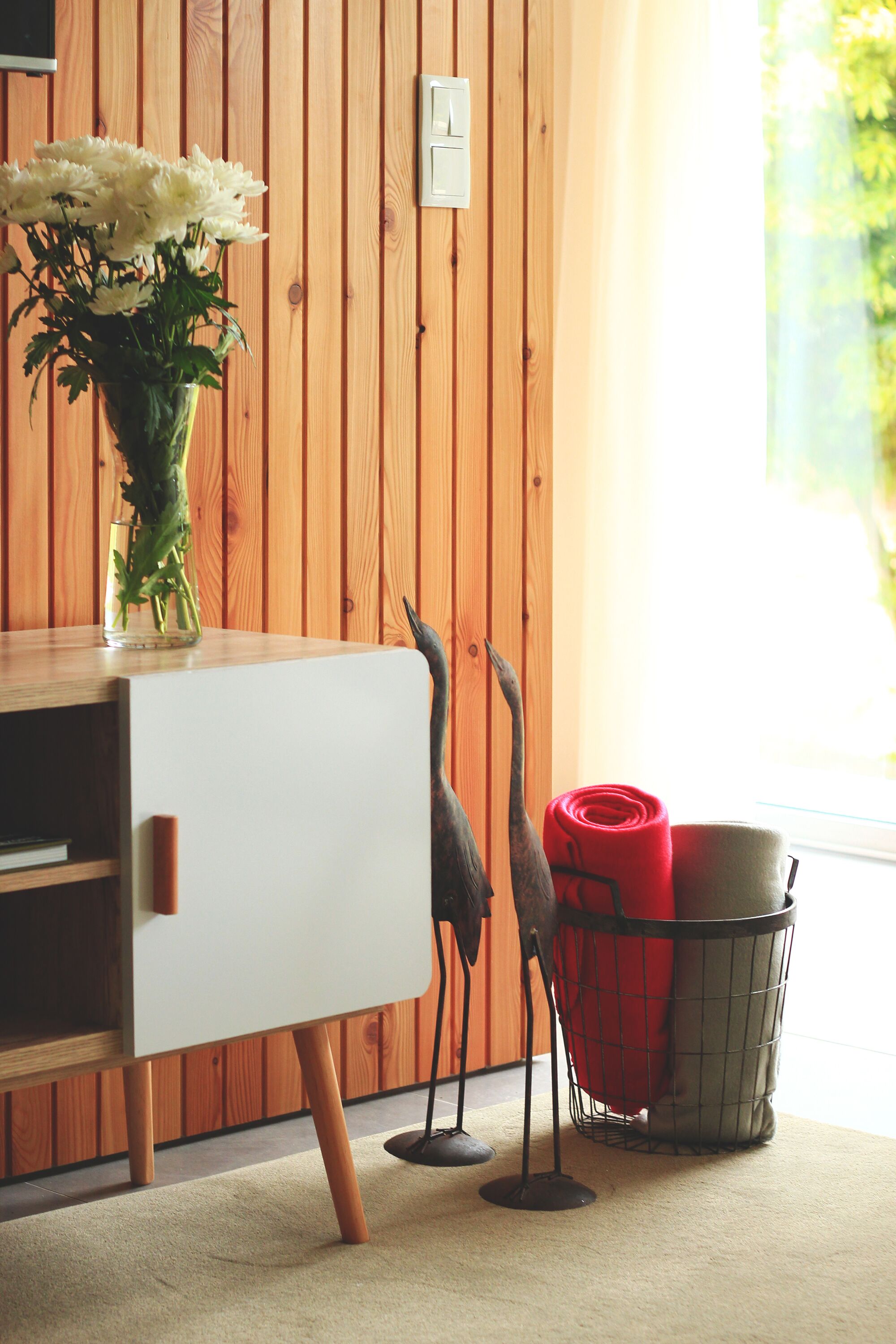 Una pared de madera, un armario blanco, un jarrón con flores, dos aves decorativas y una cesta roja y gris.