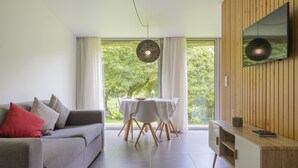 A modern living room with a grey sofa, a dining table with chairs, a wooden TV stand, and a large window with a view of greenery.