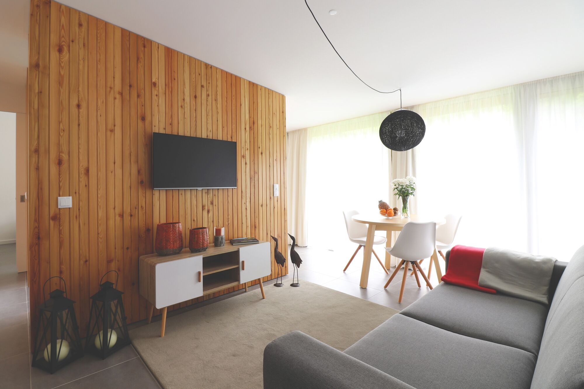 A modern living room with a wooden wall, a flat-screen TV, a dining area with a table and chairs, and a grey sofa.