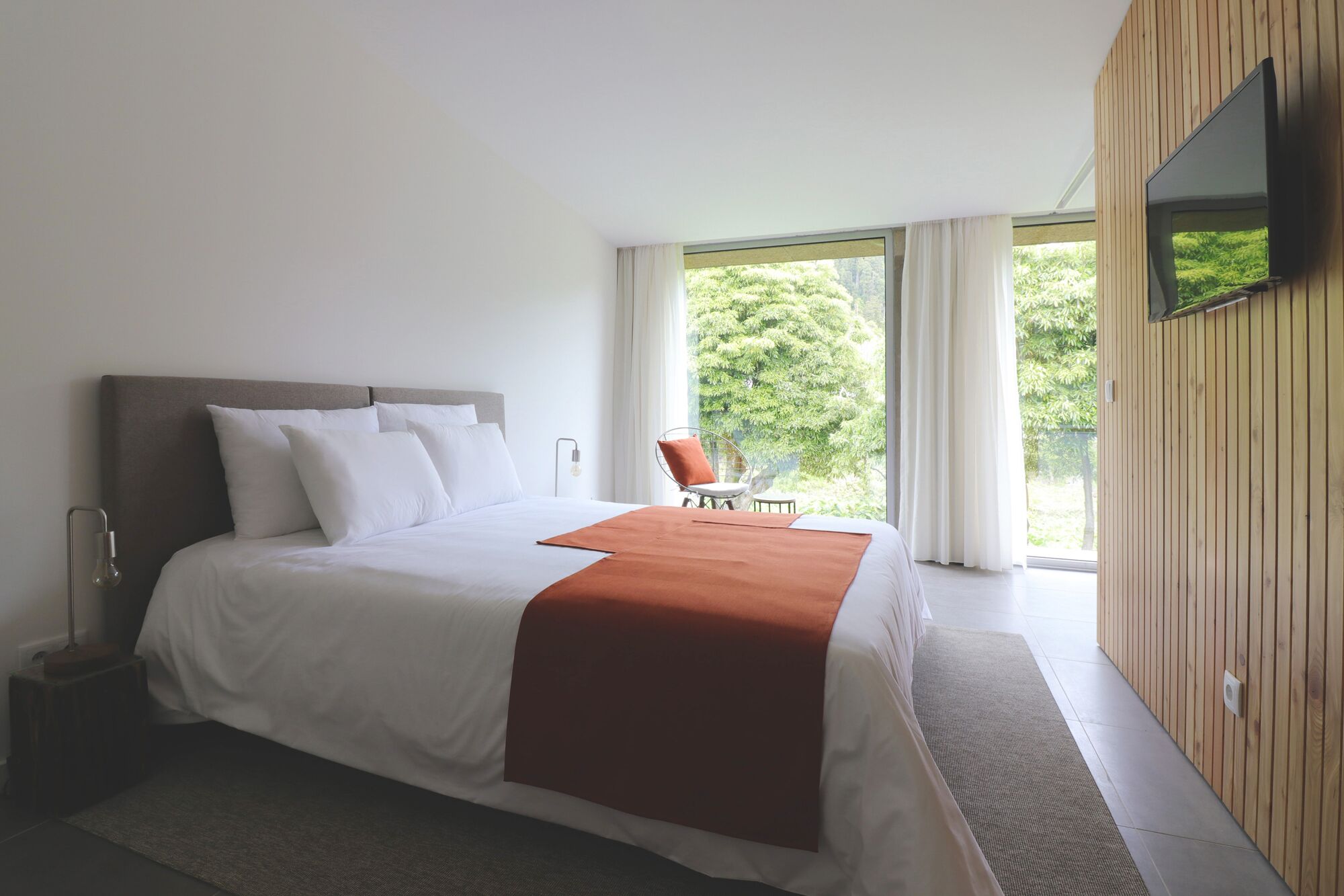 Una habitación de hotel moderna con una cama grande, un televisor montado en la pared y vistas al exterior con zonas verdes.