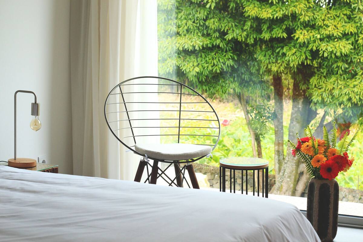 A bedroom with a bed, a chair, a small table, a lamp, and a window with a view of trees.