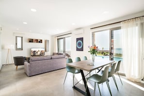 A modern living room with a sofa, dining table, and chairs, a TV, and a view of the ocean. - Beachfront Villa w/Private Pool, Kids area and BBQ (Stavromenos)