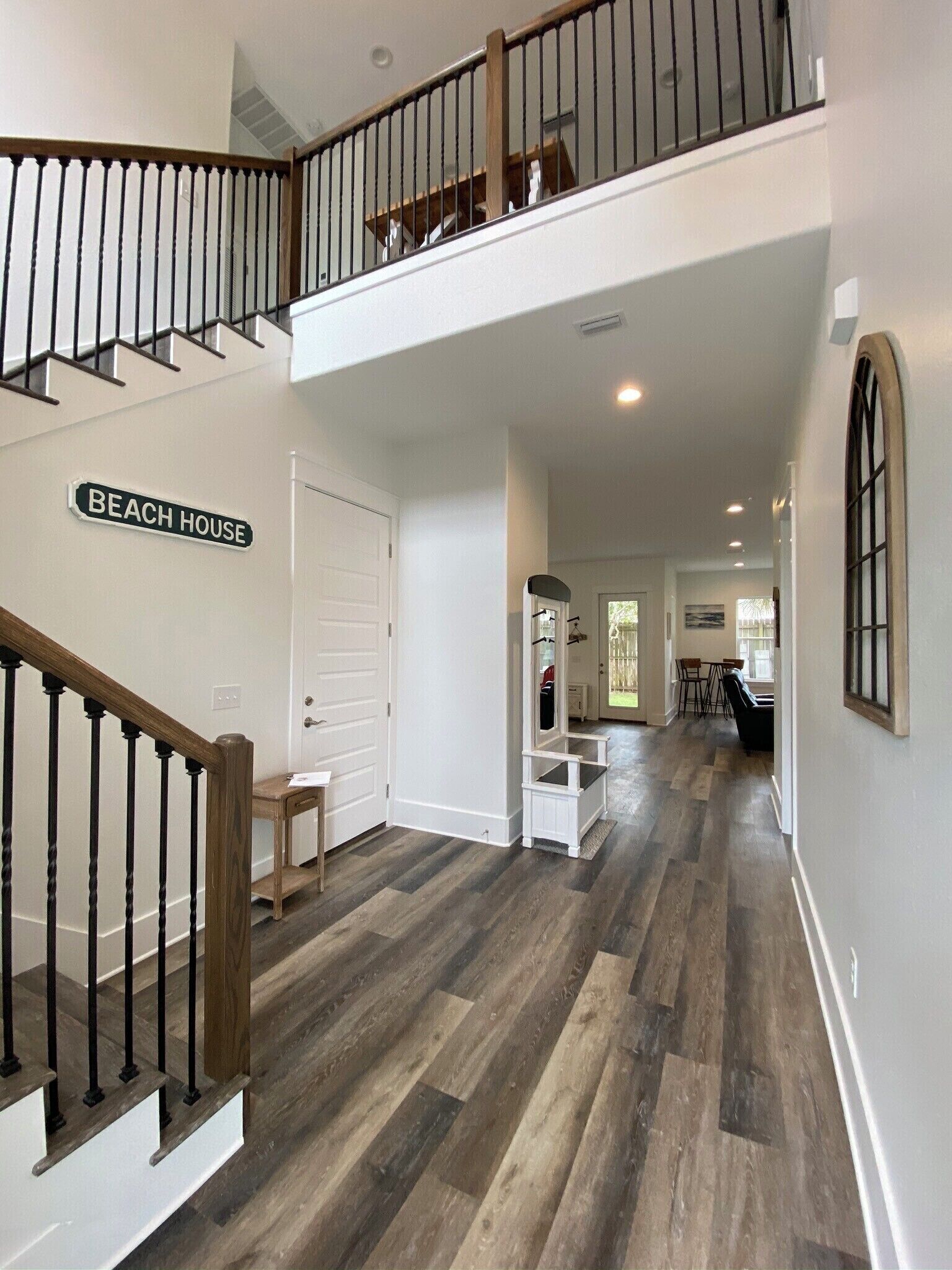 A spacious hallway with a wooden staircase leading to a second floor. A sign on the wall reads "BEACH HOUSE".