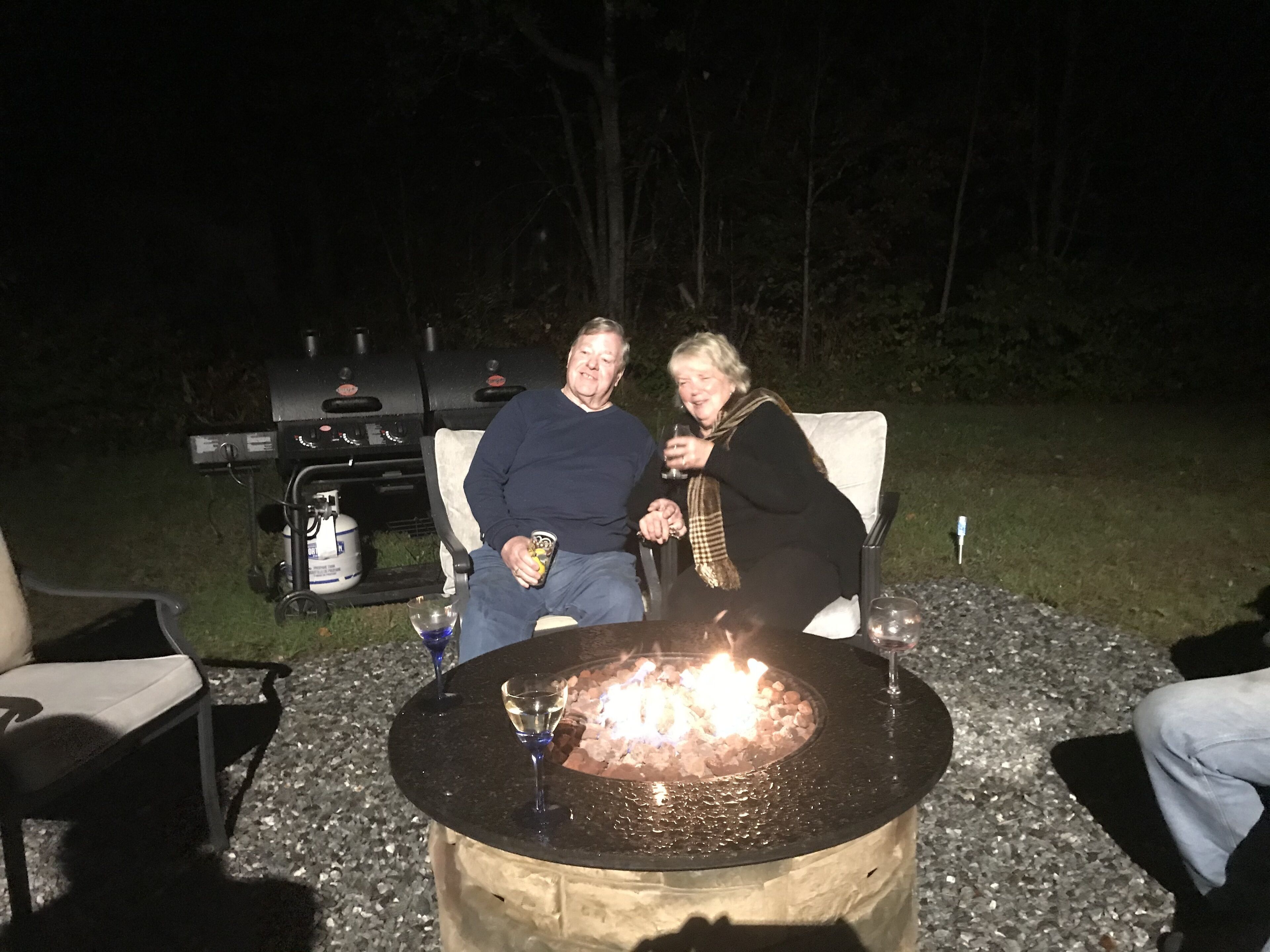 To personer sidder ved et udendørs bålsted, mens en grill står i baggrunden.