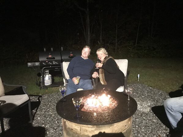 To personer sidder ved et udendørs bålsted, mens en grill står i baggrunden.