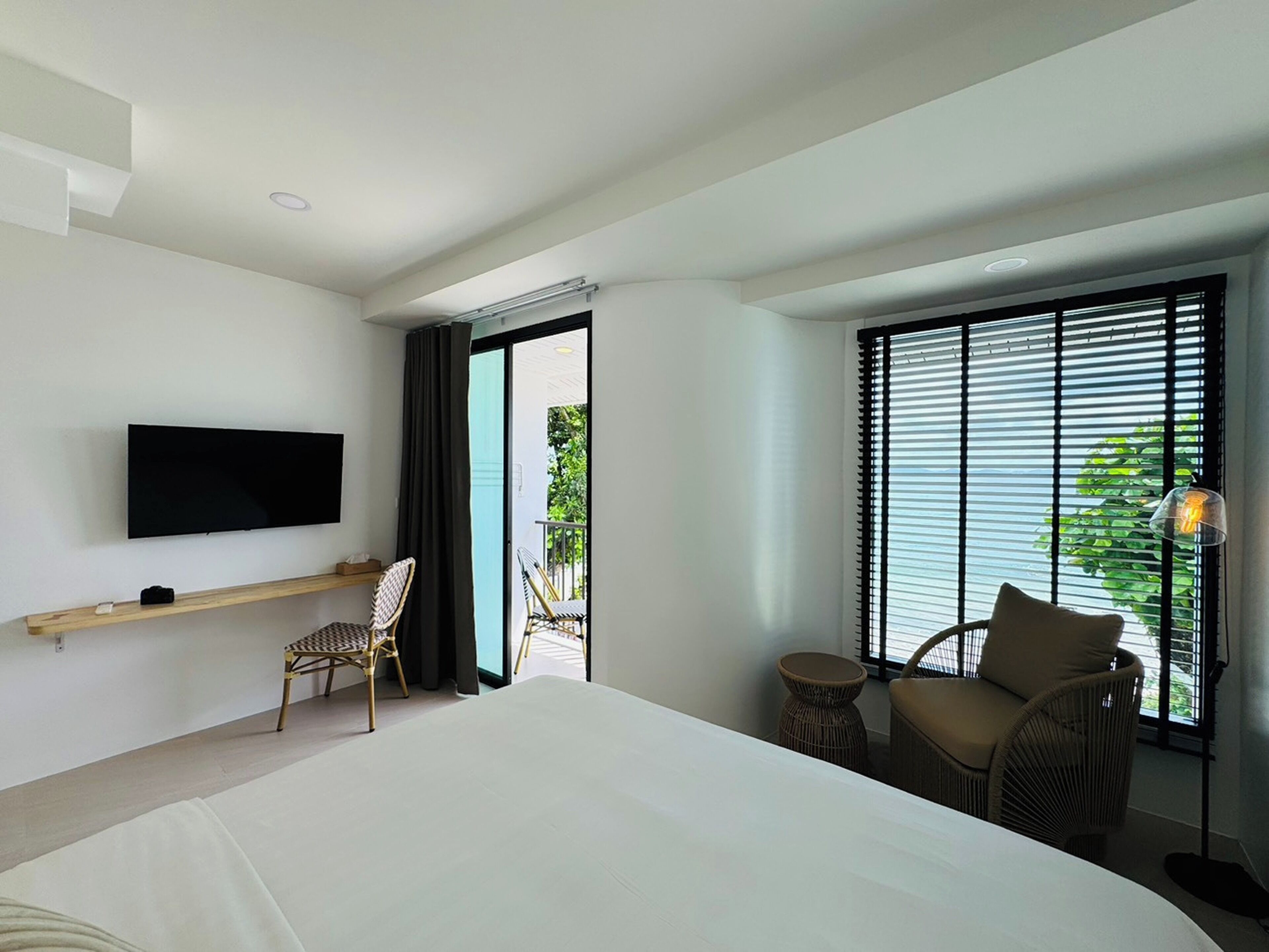 A hotel room with a bed, a TV mounted on the wall, a chair, a small table, and a window with blinds.