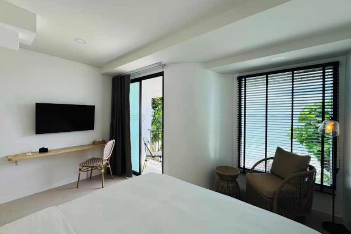 A hotel room with a bed, a TV mounted on the wall, a chair, a small table, and a window with blinds.